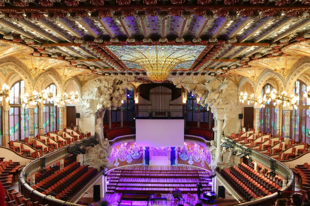 Interior view of an ornate concert hall at L’Auditori Barcelona, featuring a stained glass ceiling, sculpted walls, chandeliers, red seats, a stage, and a large screen at the center.